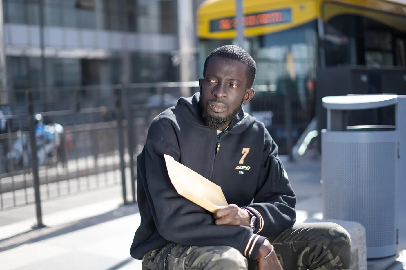 Jeune homme assis avec une enveloppe près d’un arrêt de bus, portrait KARIMKO pour le CETUD.